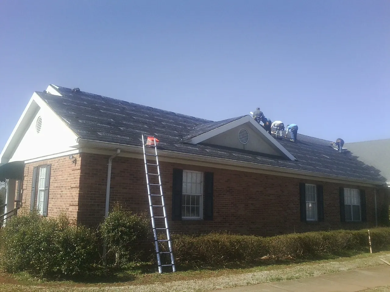 Crew tearing shingles off a brick ranch roof with a ladder set — Metal Roof Repair work by our crew in Arbon